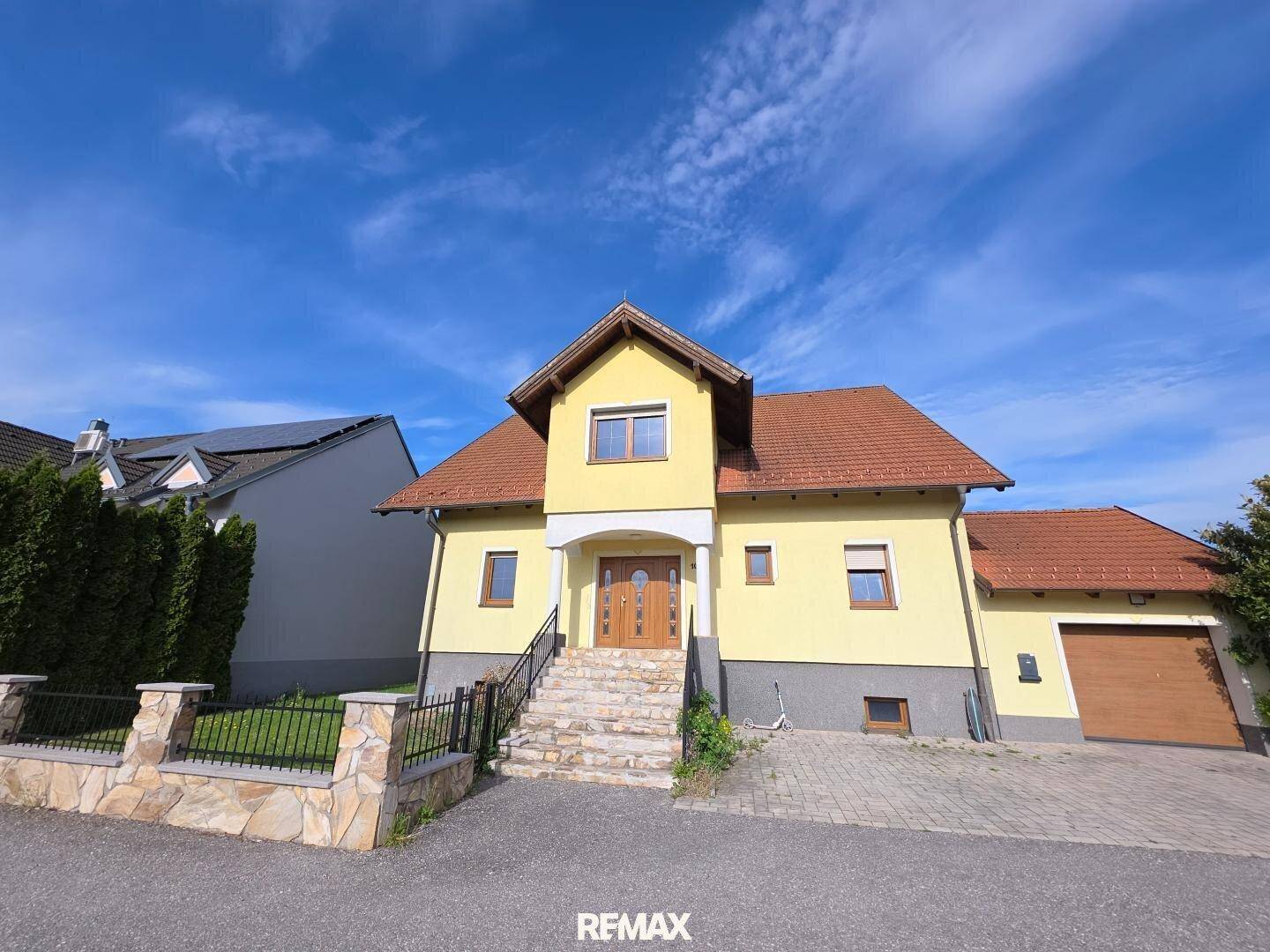 Gelbes Einfamilienhaus mit Satteldach, gepflasterter Auffahrt und Steintreppe zum Eingang.