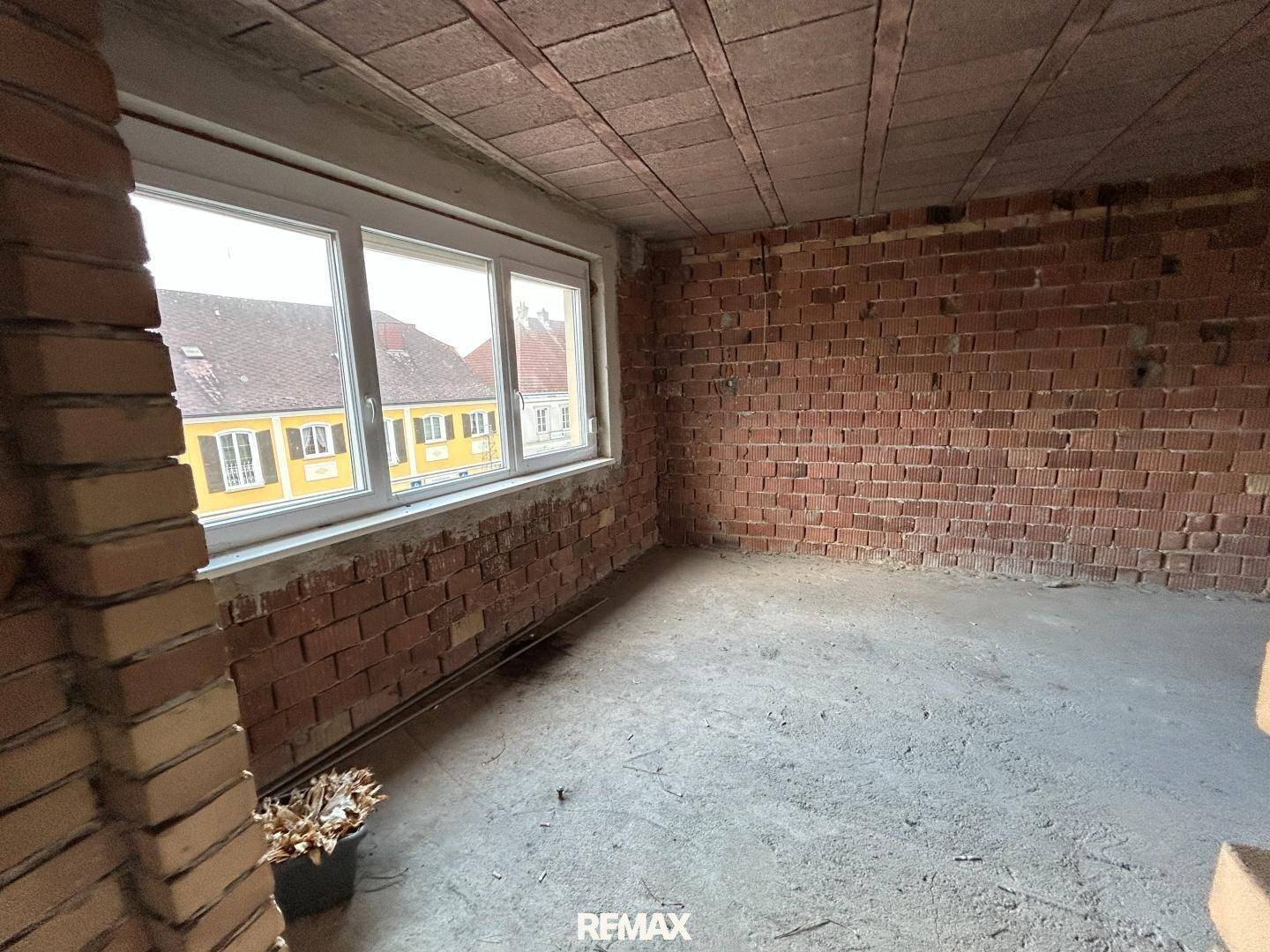 Rohbauzimmer mit unverputzten Ziegelwänden, Betonboden und einem großen Fenster zur Straße.