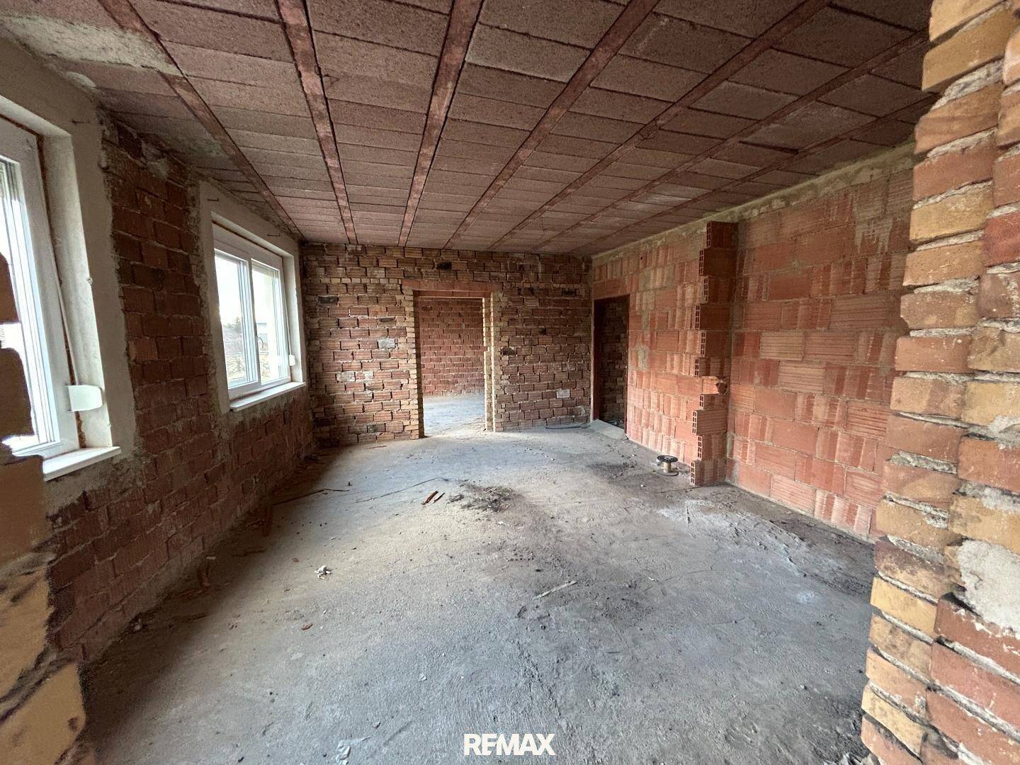 Rohbauzimmer mit unverputzten Ziegelwänden, Betonboden und Fenstern, bereit für den Ausbau.