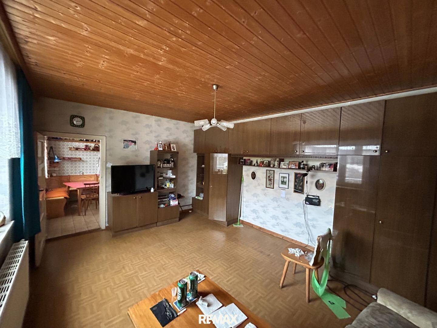 Wohnzimmer mit Holzdecke, Parkettboden und Blick in die angrenzende Küche.