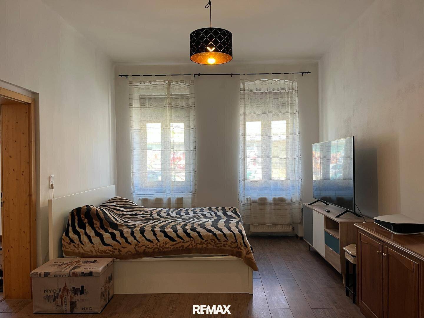 Schlafzimmer mit Bett, Fernseher und zwei Fenstern zur Straßenseite, einfach eingerichtet.