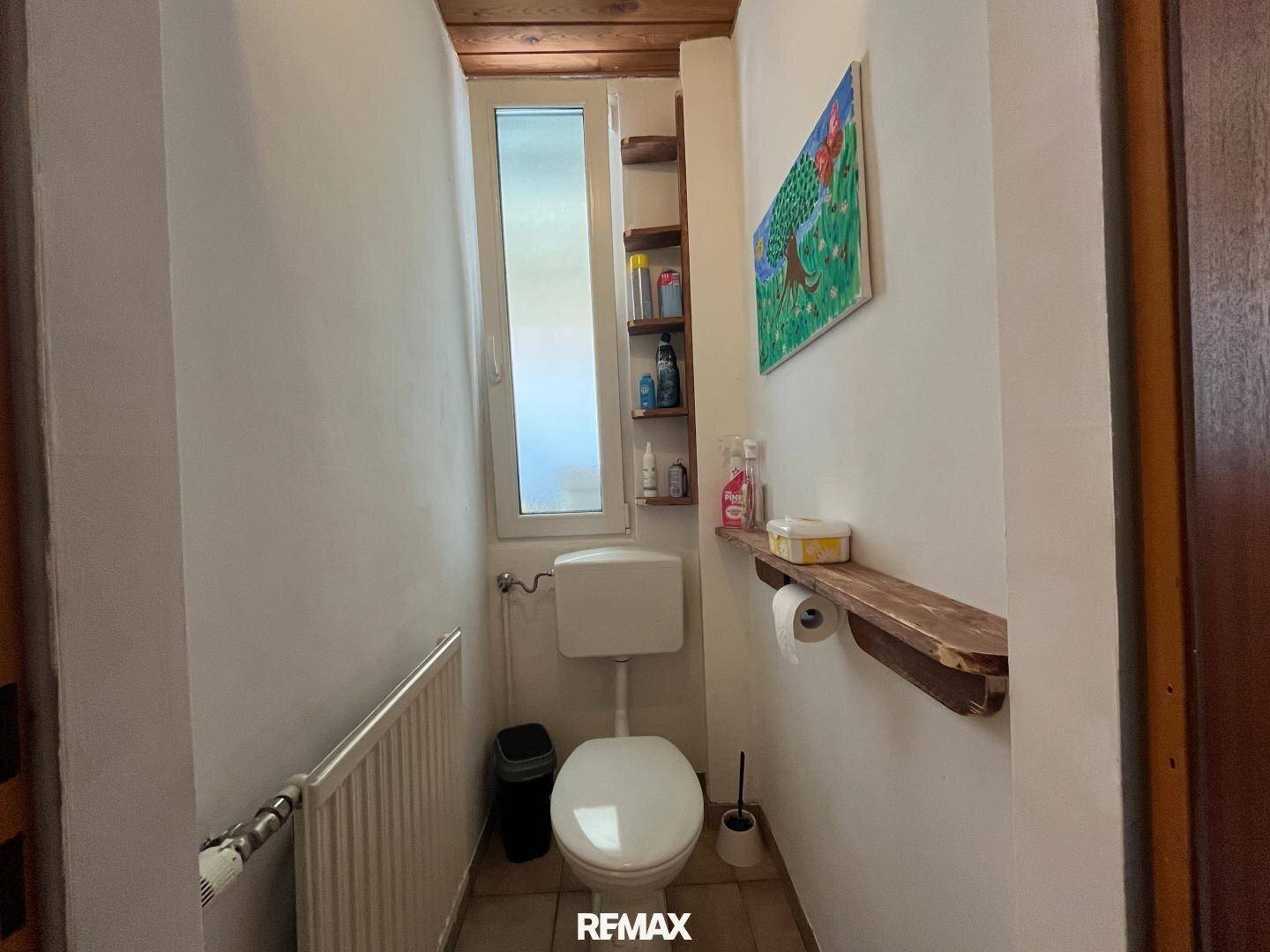 Kleines WC mit Fenster, Toilette und Holzregal, einfach und zweckmäßig.