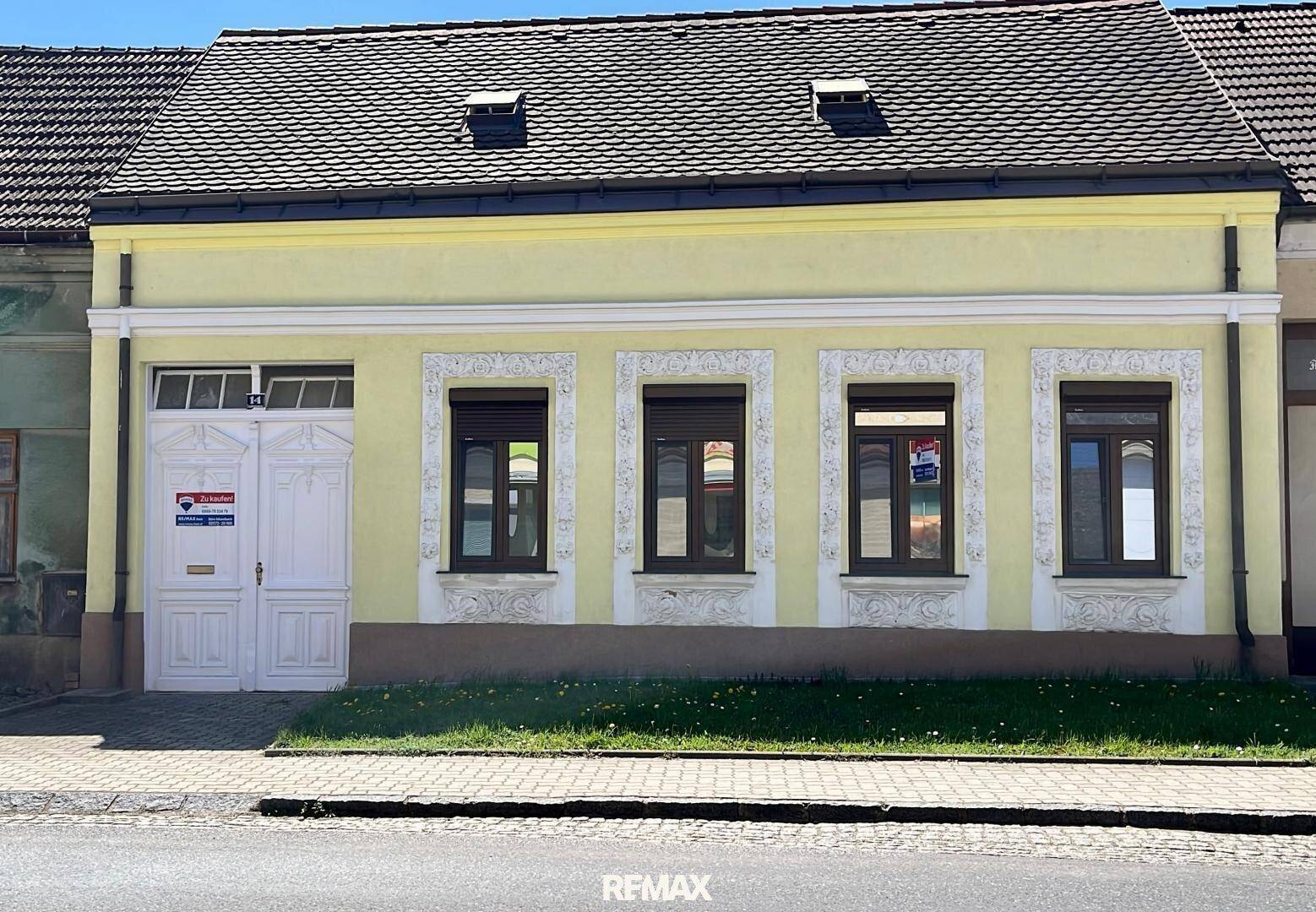 Gelbe Hausfassade mit braunen Fenstern und dekorativen Stuckelementen unter einem Satteldach.