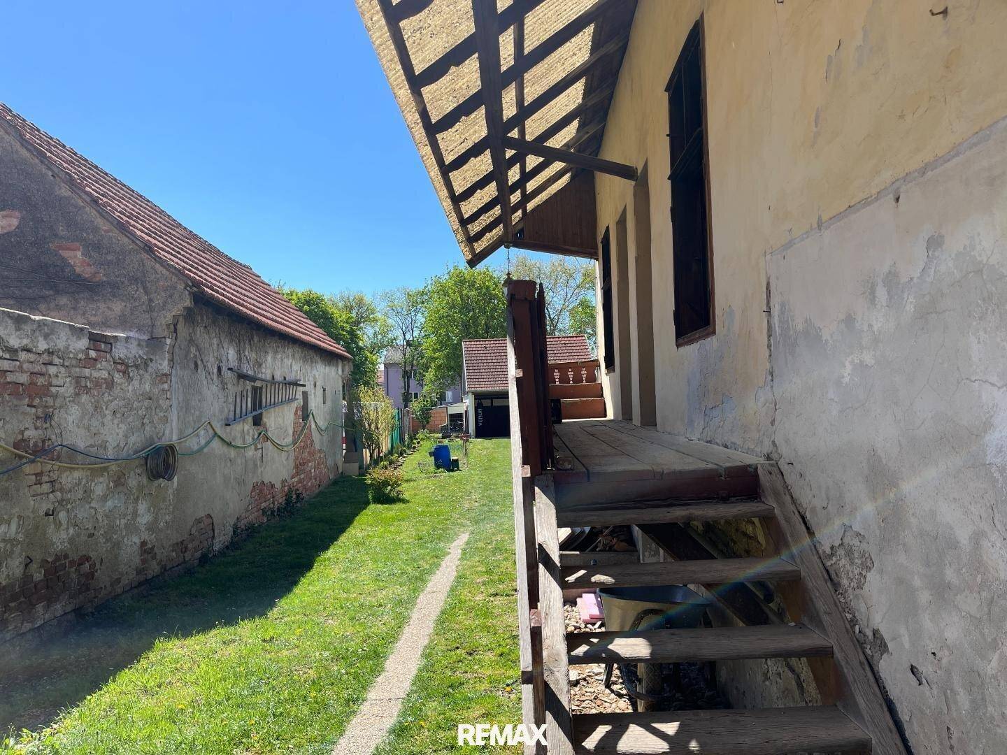 Grüner Gartenbereich mit alter Hausfassade und Holztreppe, sonnig und weitläufig.