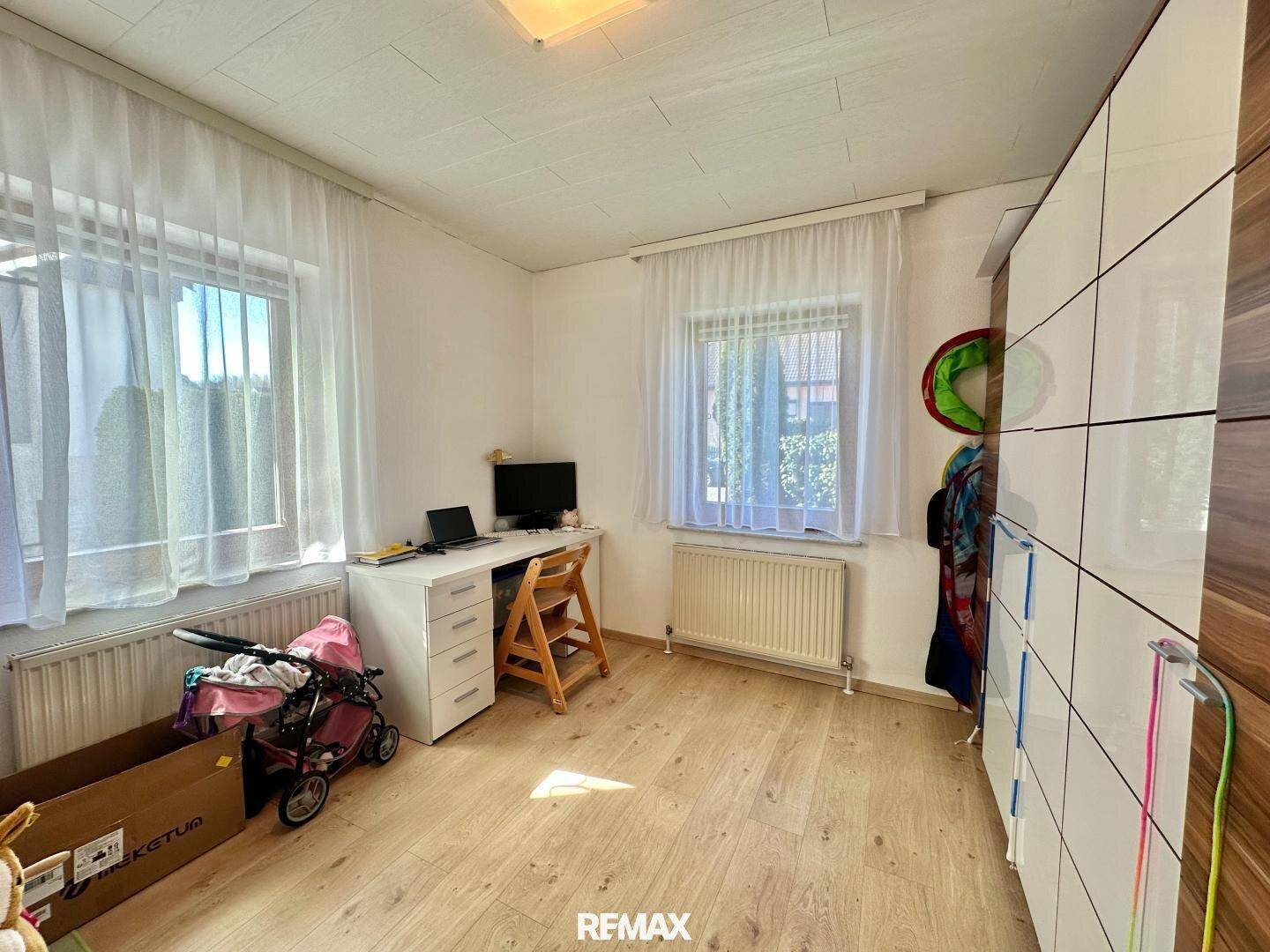 Helles Kinderzimmer mit Schreibtisch, Schrank und zwei Fenstern.