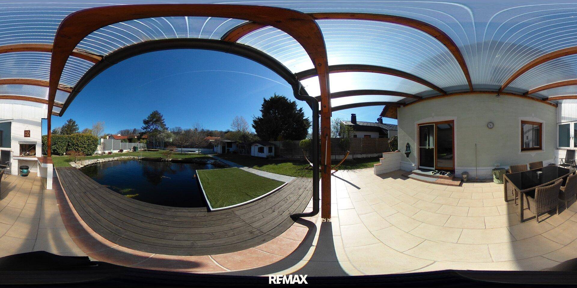 Überdachte Terrasse mit Sitzgelegenheiten und Blick auf den Garten mit Teich an einem sonnigen Tag.