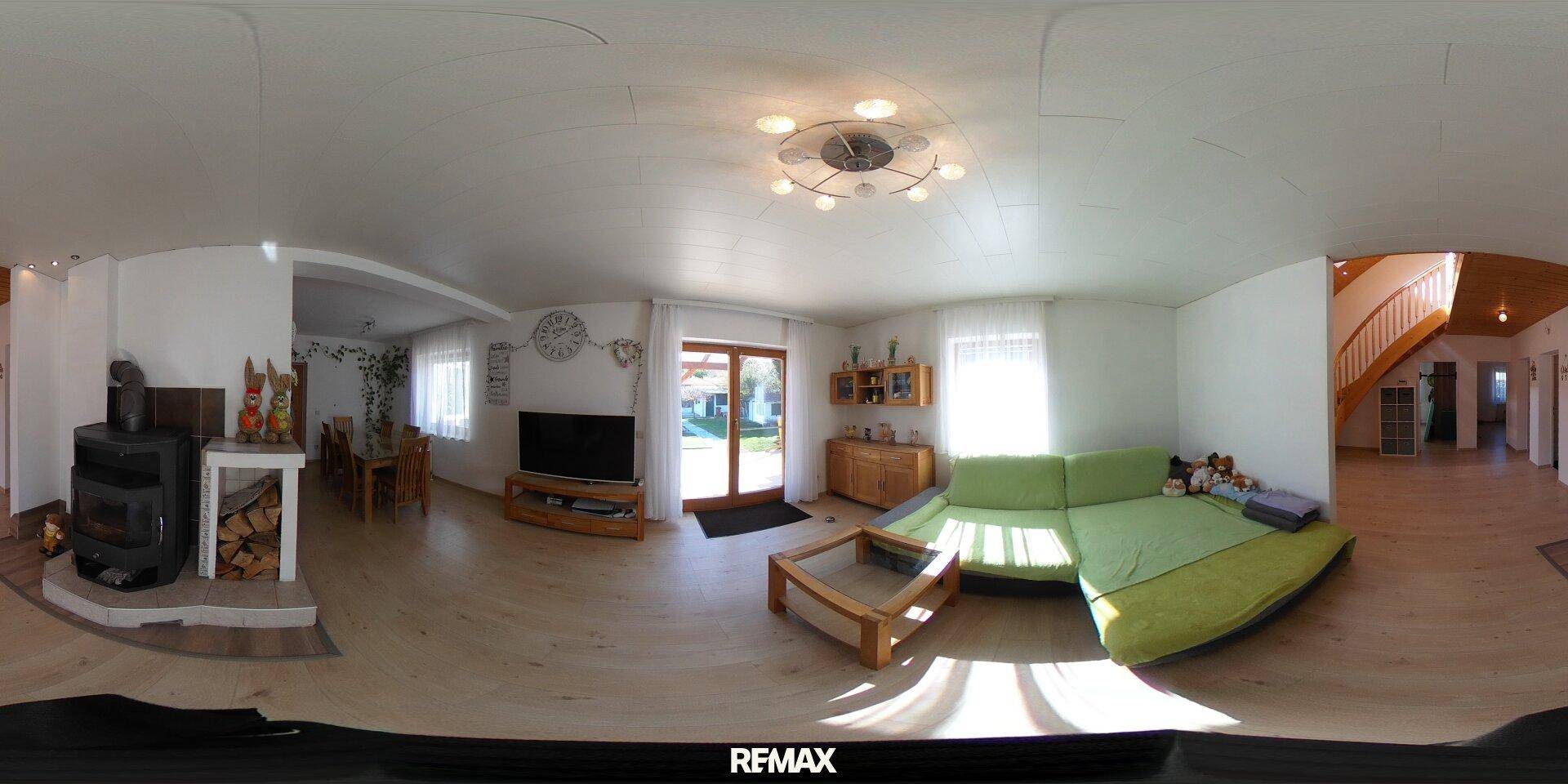 Gemütliches Wohnzimmer mit grünem Sofa, Fernseher und einem Holzofen, hell durch große Fenster.