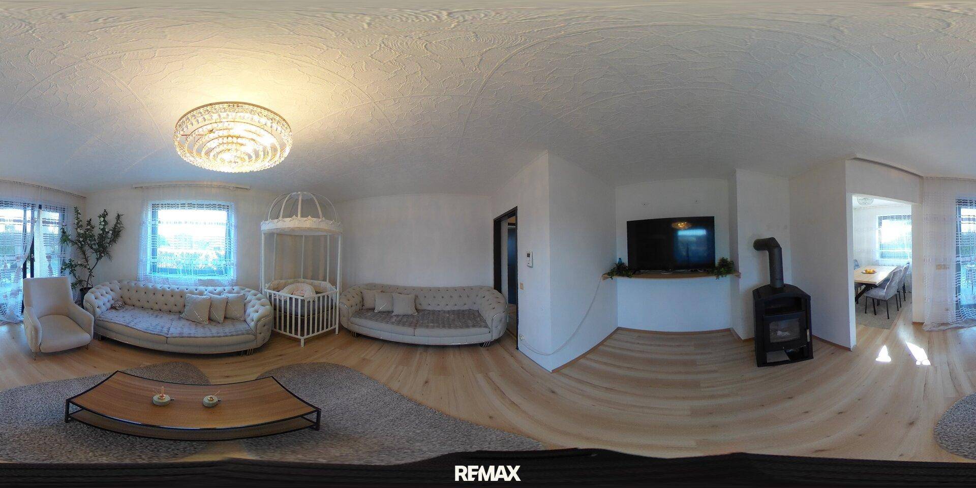 Panoramablick auf ein gemütliches Wohnzimmer mit Sofa, Kaminofen und Zugang nach draußen.