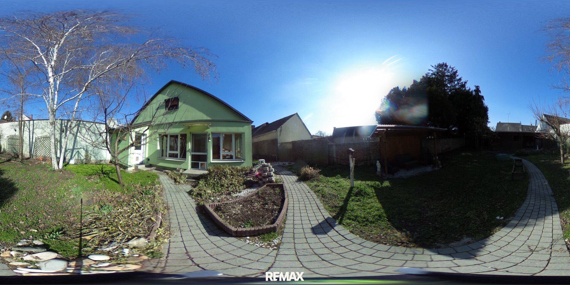 Grünes Haus mit gepflasterten Wegen und einem sonnigen Gartenbereich, umgeben von Bäumen.