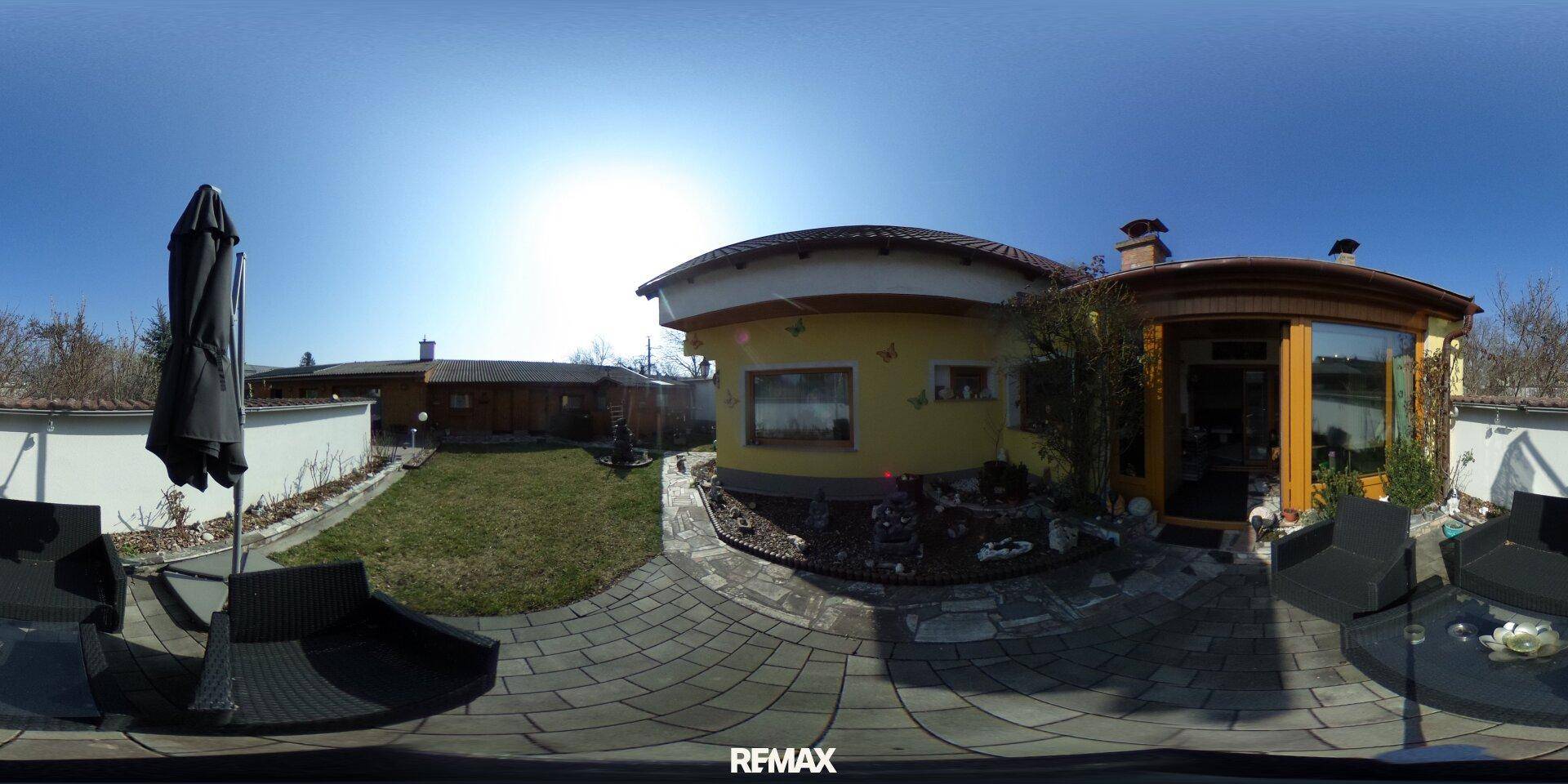 Sonniger Gartenbereich mit gepflasterter Terrasse, Sitzgelegenheiten und Rasenfläche hinter dem Haus.