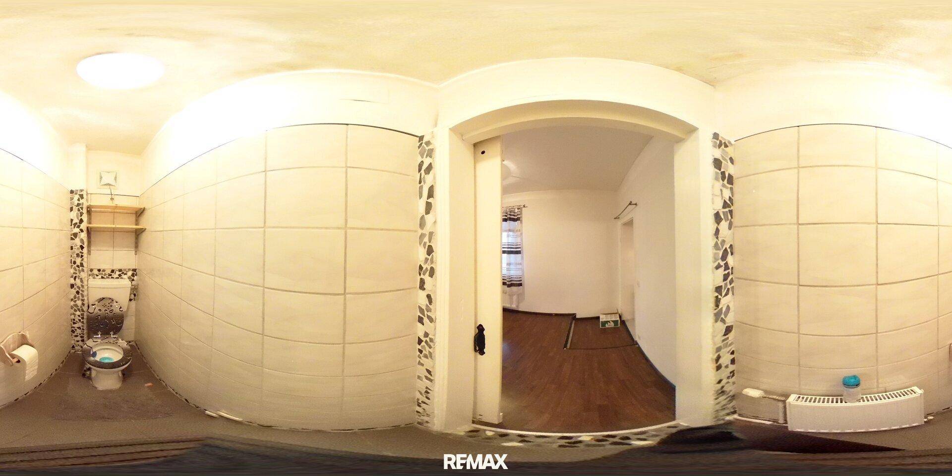 Panoramaaufnahme einer Toilette mit weißen Fliesen und einem kleinen Waschbecken.