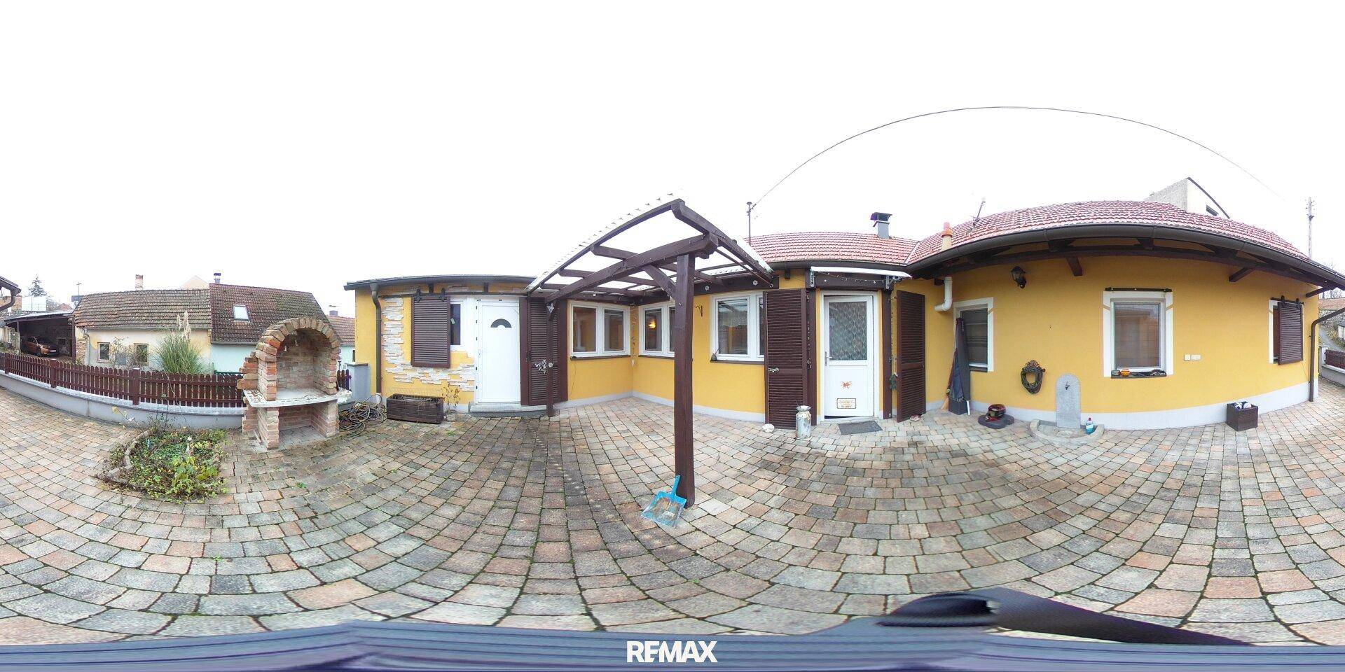 Gepflasterter Innenhof mit gelber Hausfassade, Grillplatz und einer Holzpergola für Schatten.