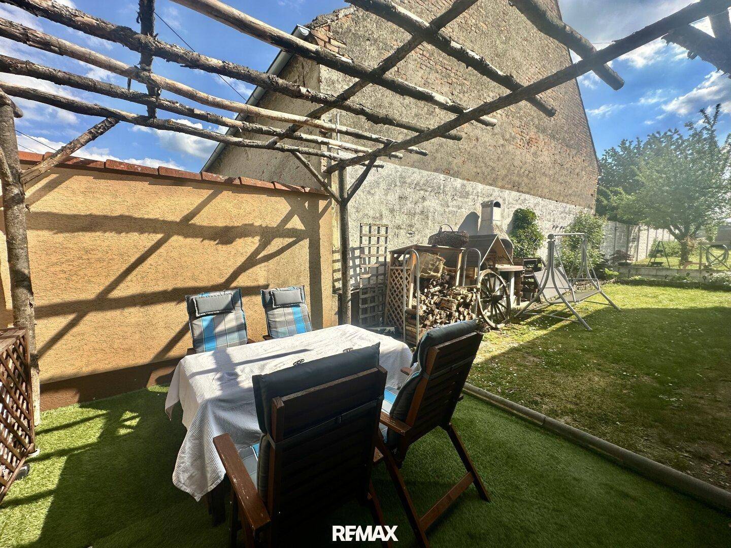 Überdachte Terrasse mit Esstisch und Stühlen, Kunstrasen und Blick auf den Gartenbereich.