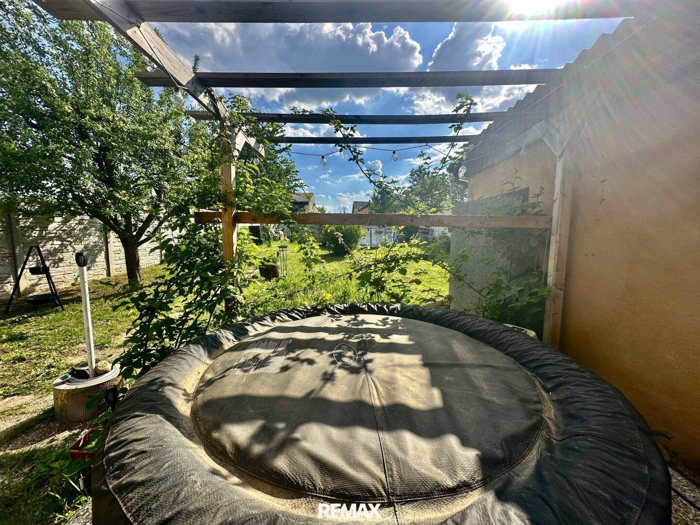 Überdachter Whirlpool im Garten unter einer Holzpergola an einem sonnigen Tag mit viel Grün.