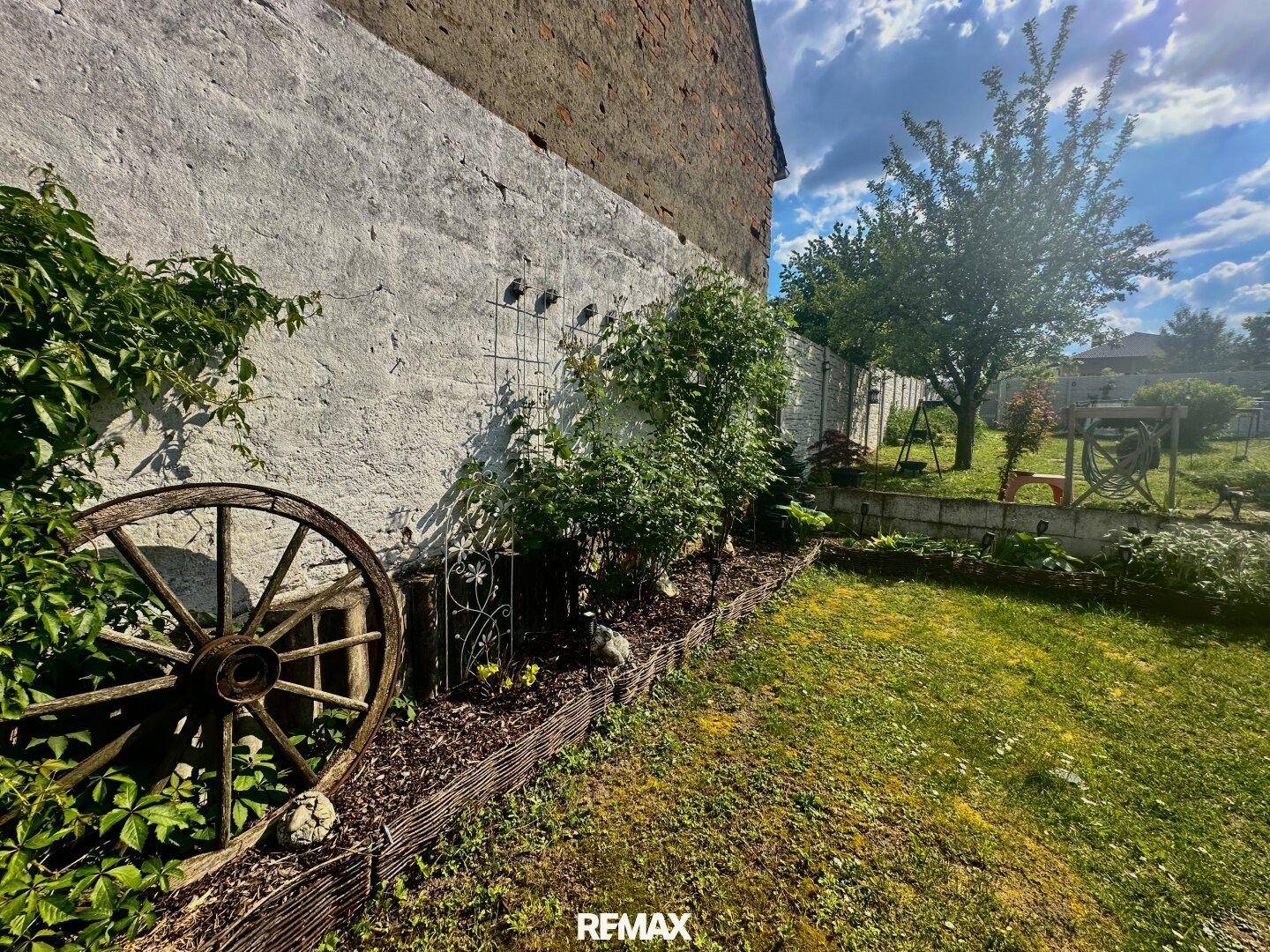 Gartenbereich mit dekorativem Wagenrad, bepflanztem Beet und einer rauen Hauswand im Hintergrund.