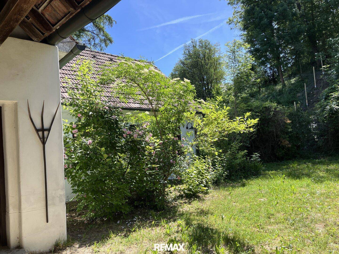 Seitlicher Blick auf das Landhaus mit blühenden Büschen und einem gepflegten Rasen unter blauem Himmel.