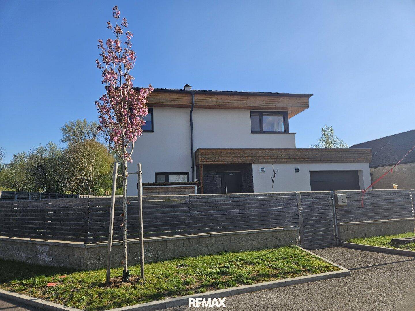 Ansicht des modernen Hauses von der Straße mit blühendem Baum und Holzzaun im Sonnenschein.