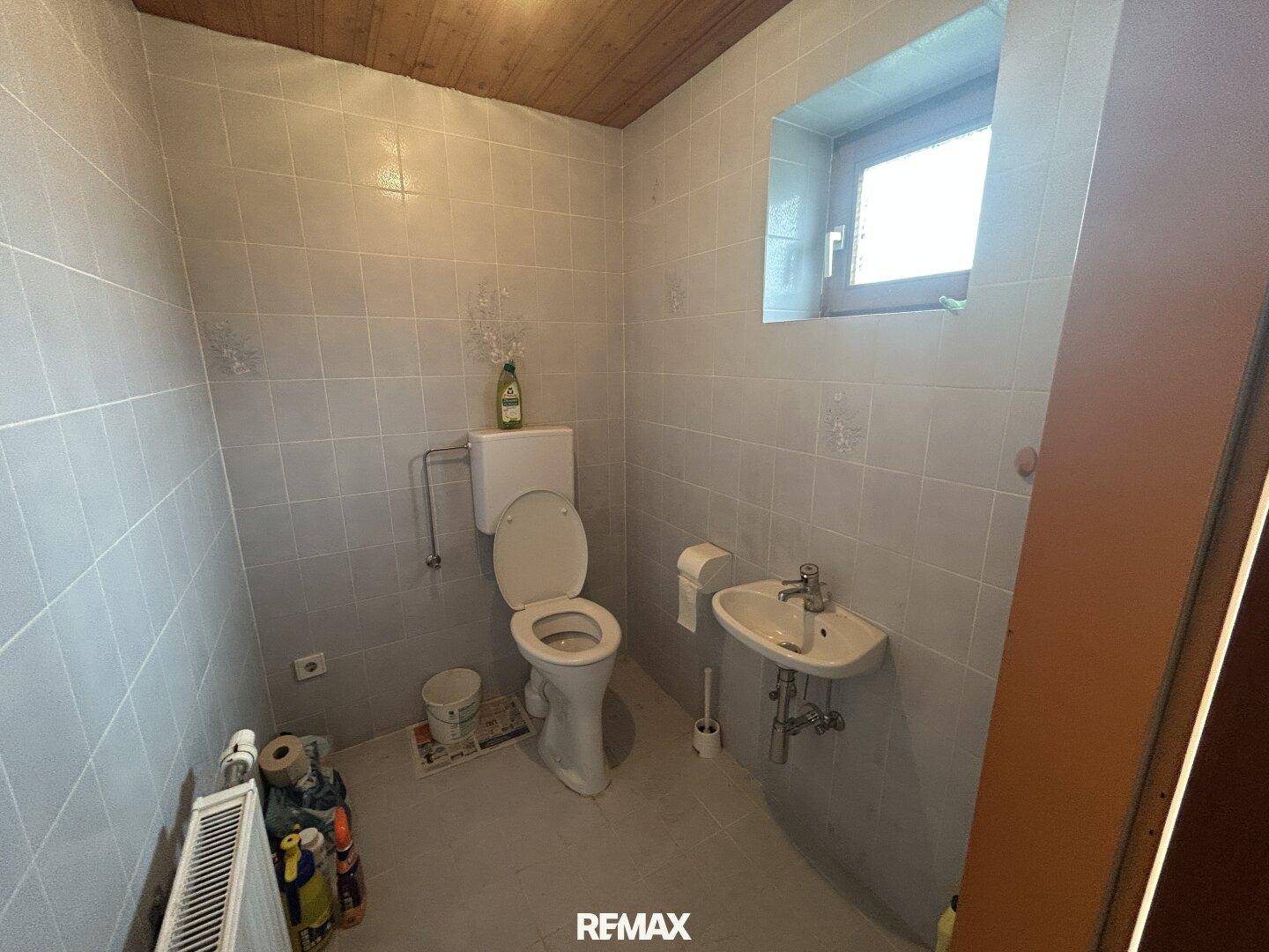 Kleines WC mit Fliesenboden, Toilette, Waschbecken und einem Fenster für Tageslicht.