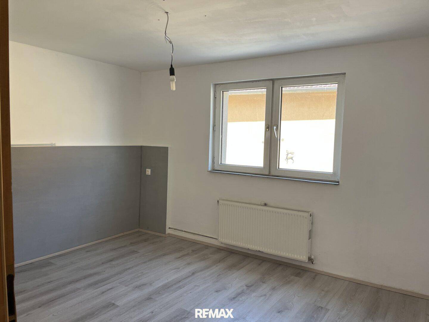 Leerer Raum mit Laminatboden, Heizkörper und einem Fenster, das viel Licht hereinlässt.