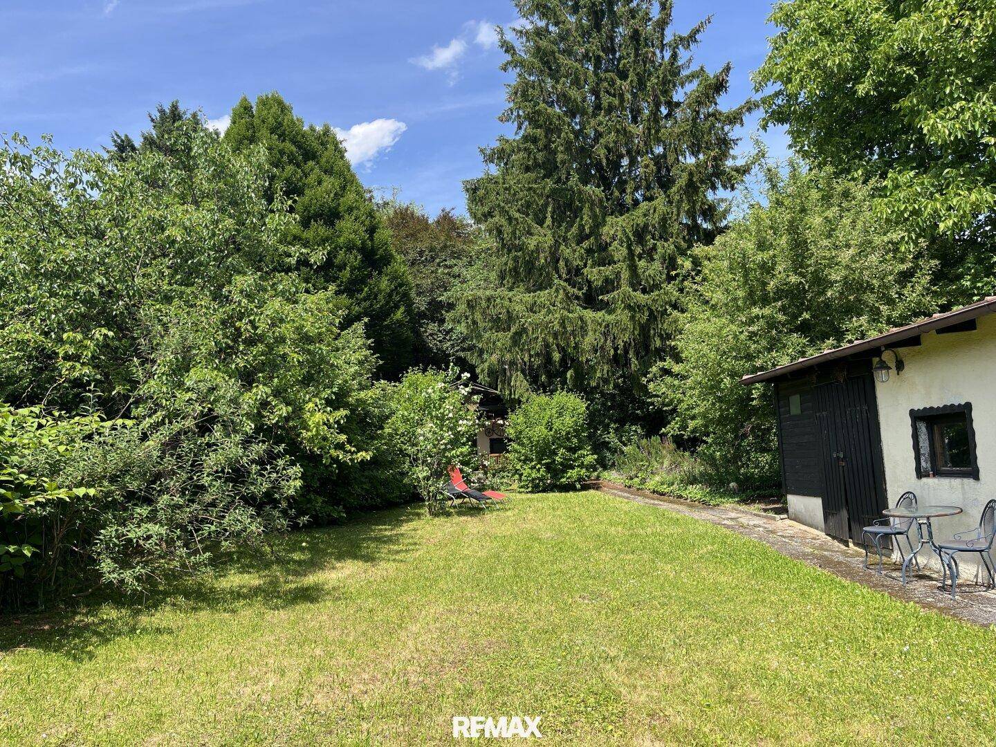 Großer, gepflegter Garten mit Rasenfläche, hohen Bäumen und einem kleinen Nebengebäude.