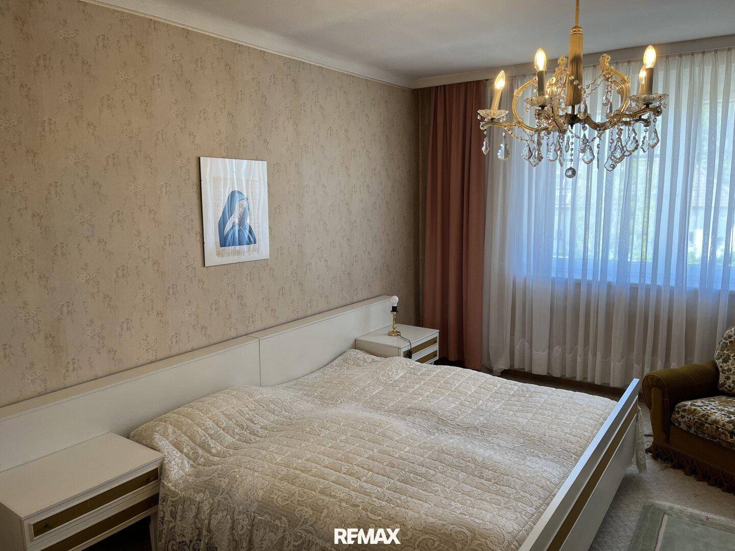 Schlafzimmer mit Doppelbett, gemusterter Tapete und Kronleuchter, hell durch große Fenster.