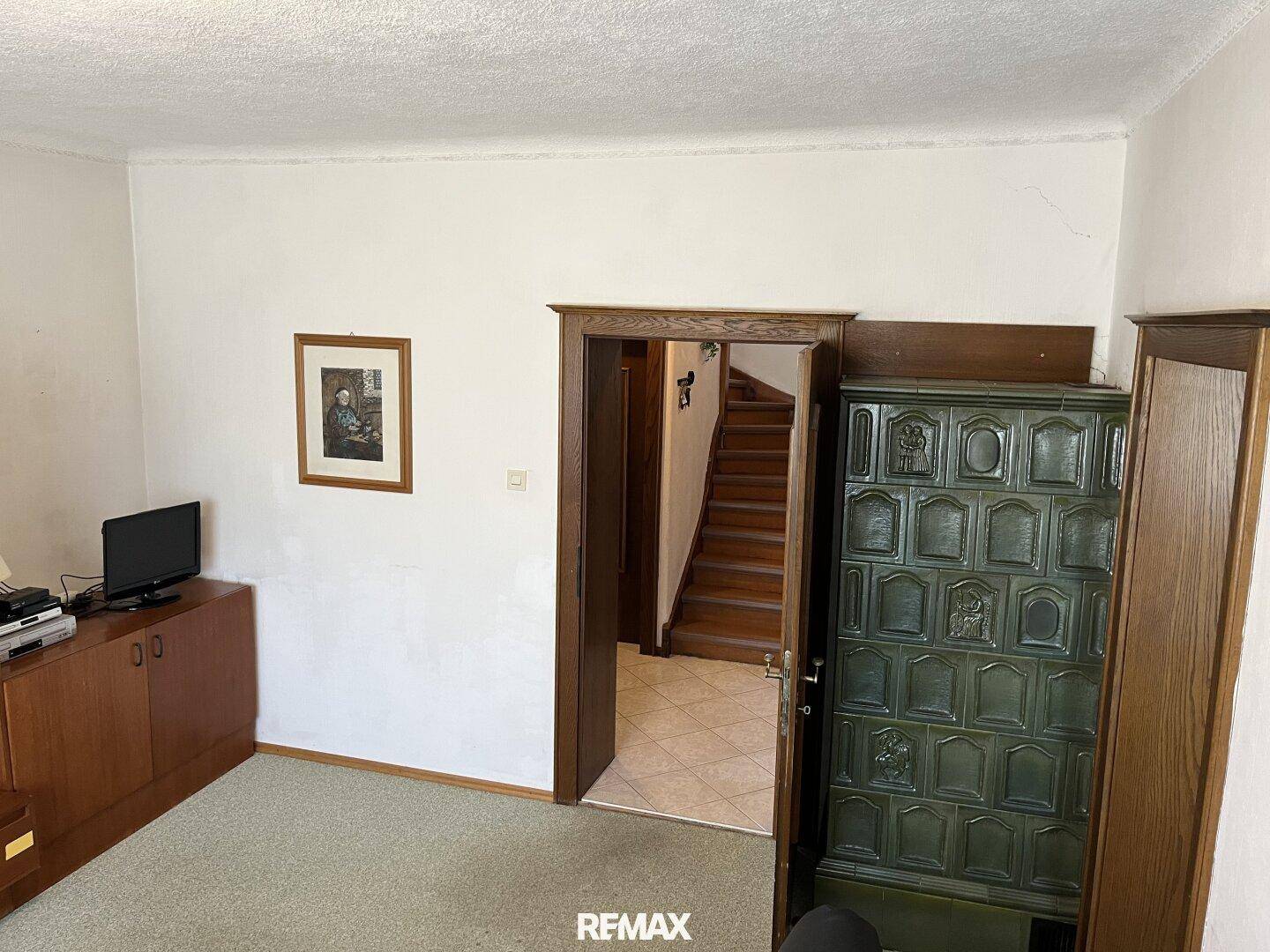 Wohnraum mit grünem Kachelofen, Teppichboden und Zugang zur Holztreppe.