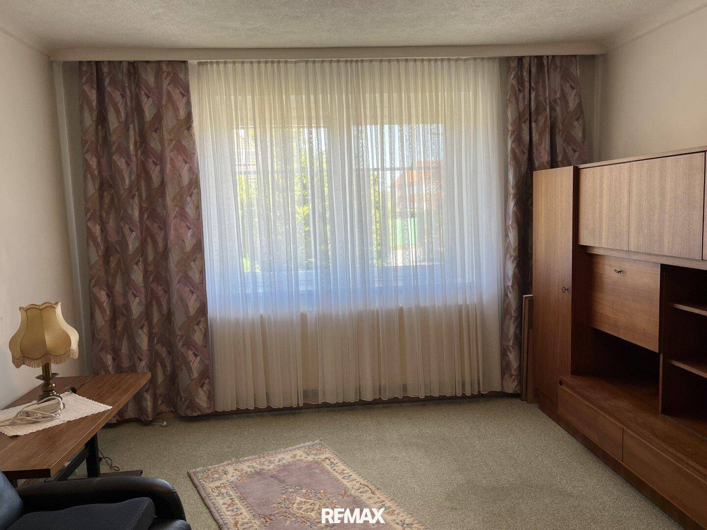 Helles Wohnzimmer mit großem Fenster, Teppichboden und traditionellen Vorhängen.