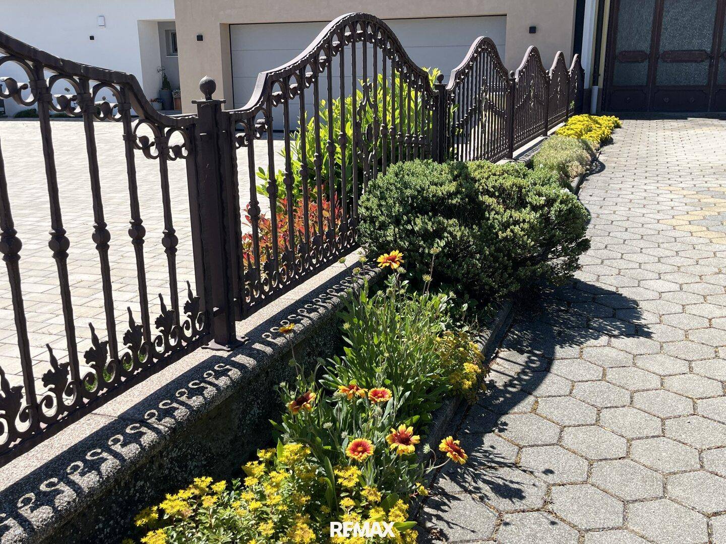 Dekorativer Metallzaun mit bunten Blumenbeeten vor einer Garageneinfahrt.