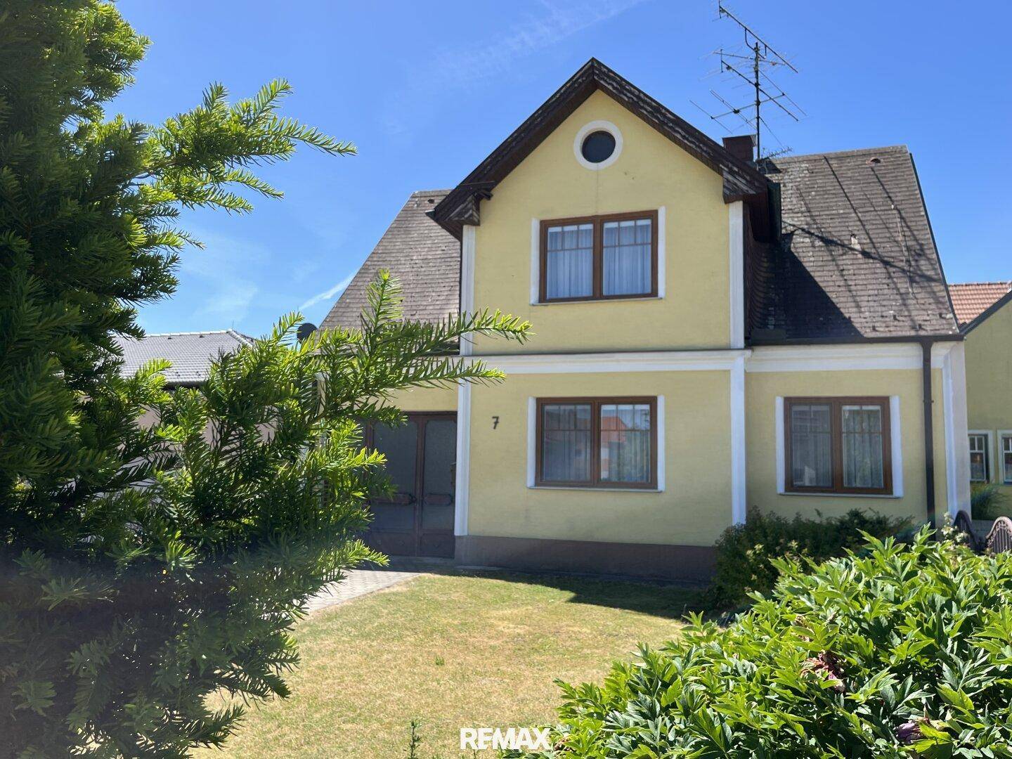 Vorderansicht des gelben Hauses, teilweise von einem grünen Busch verdeckt, unter blauem Himmel.