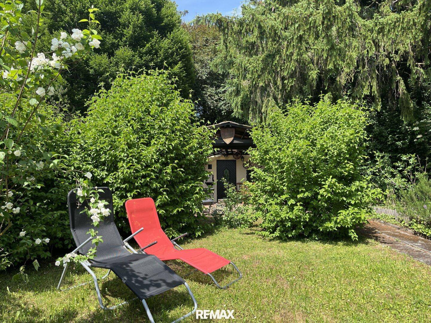 Sonnige Gartenecke mit Liegestühlen, üppigem Grün und blühenden Sträuchern.