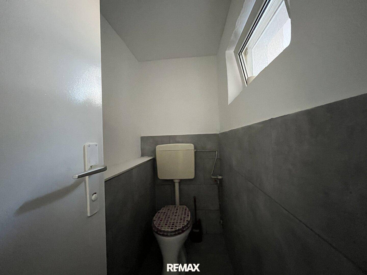 Kleines WC mit Fenster, grauen Fliesen an der Wand und einer einfachen Toilette.
