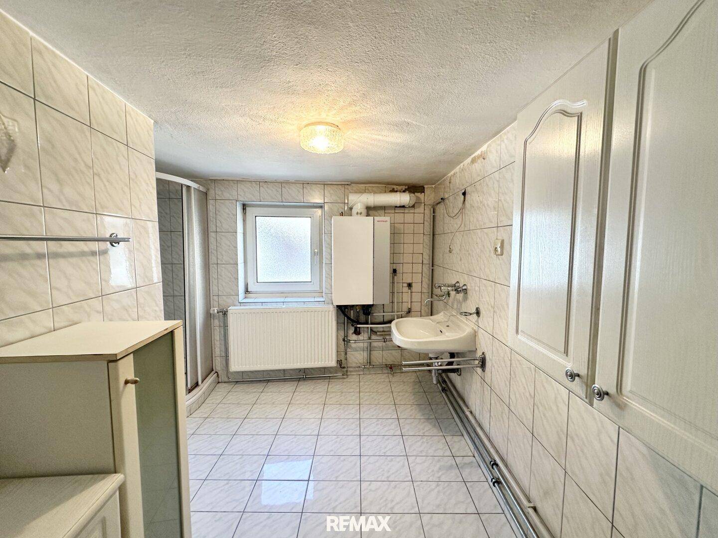 Gefliestes Badezimmer mit Dusche, Waschbecken und Heizungsanlage, in originalem Zustand.