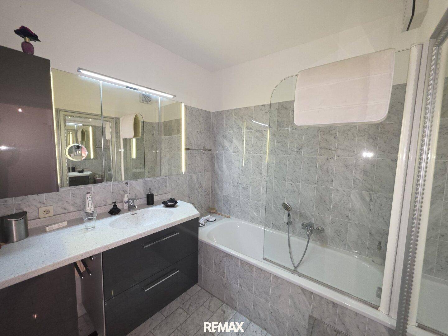 Badezimmer mit grauen Fliesen, Badewanne mit Duschwand und modernem Waschtisch mit Spiegel.
