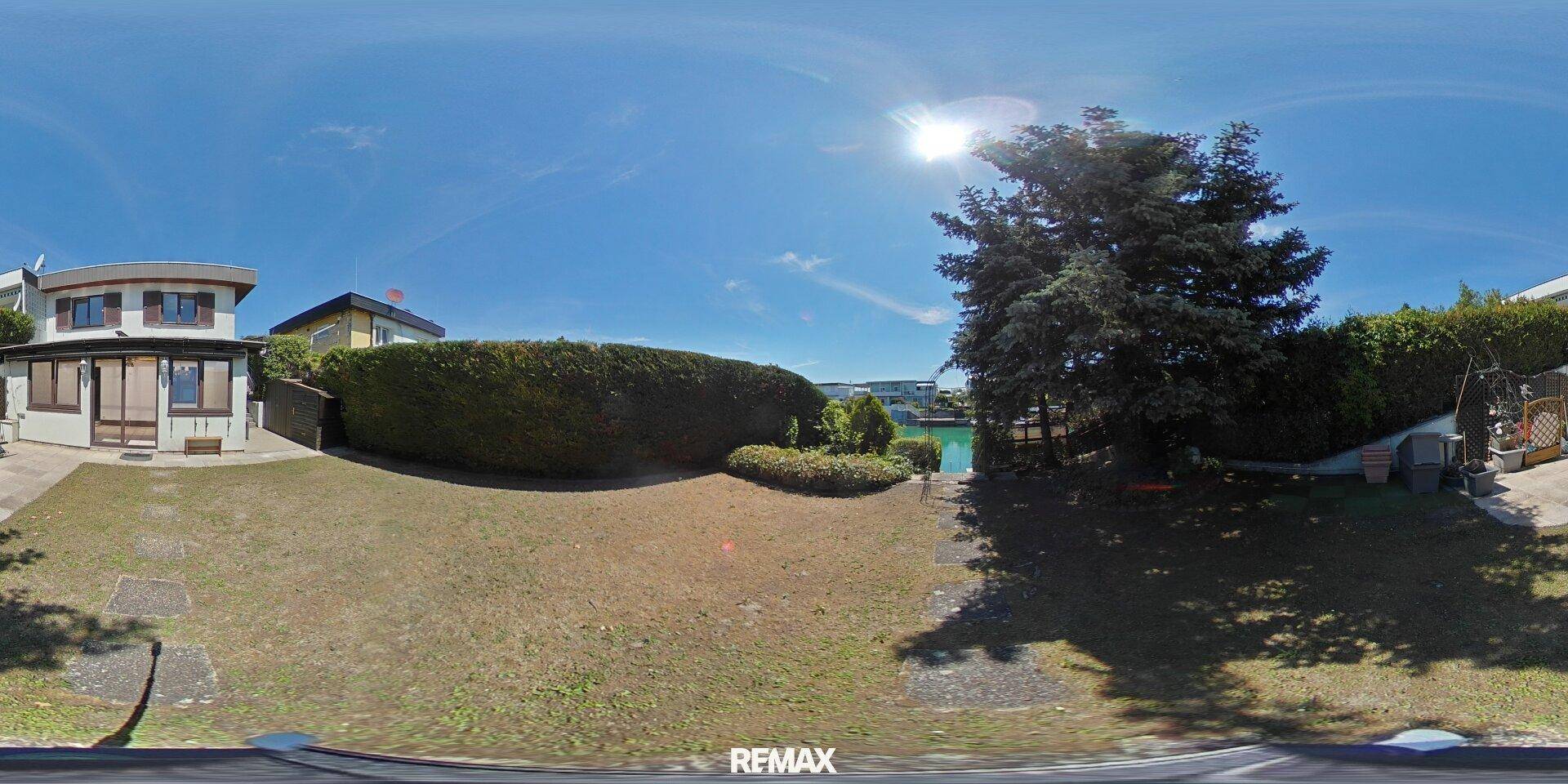 Weitläufiger Gartenbereich mit Rasen, Hecken und dem Haus im Hintergrund unter blauem Himmel.