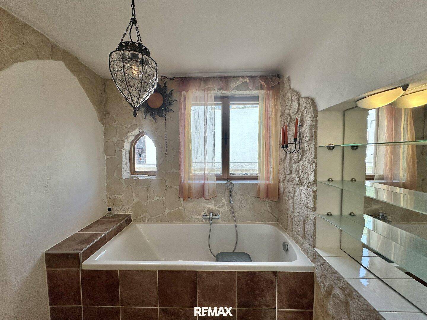 Badezimmer mit eingebauter Badewanne, gefliesten Wänden und einem kleinen Fenster für Tageslicht.