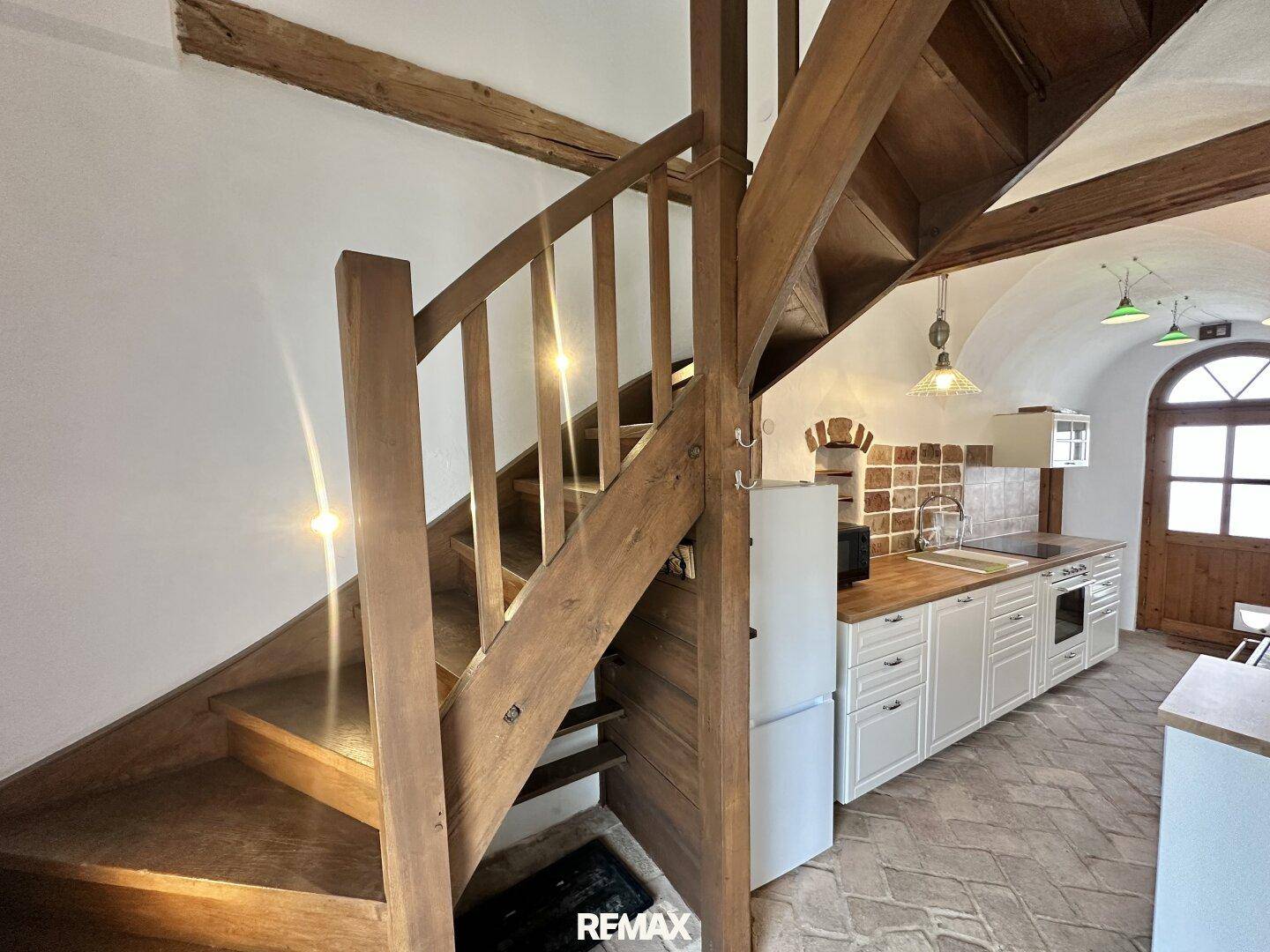 Holztreppe führt in die oberen Stockwerke, mit Blick auf die rustikale Küche im Erdgeschoss.