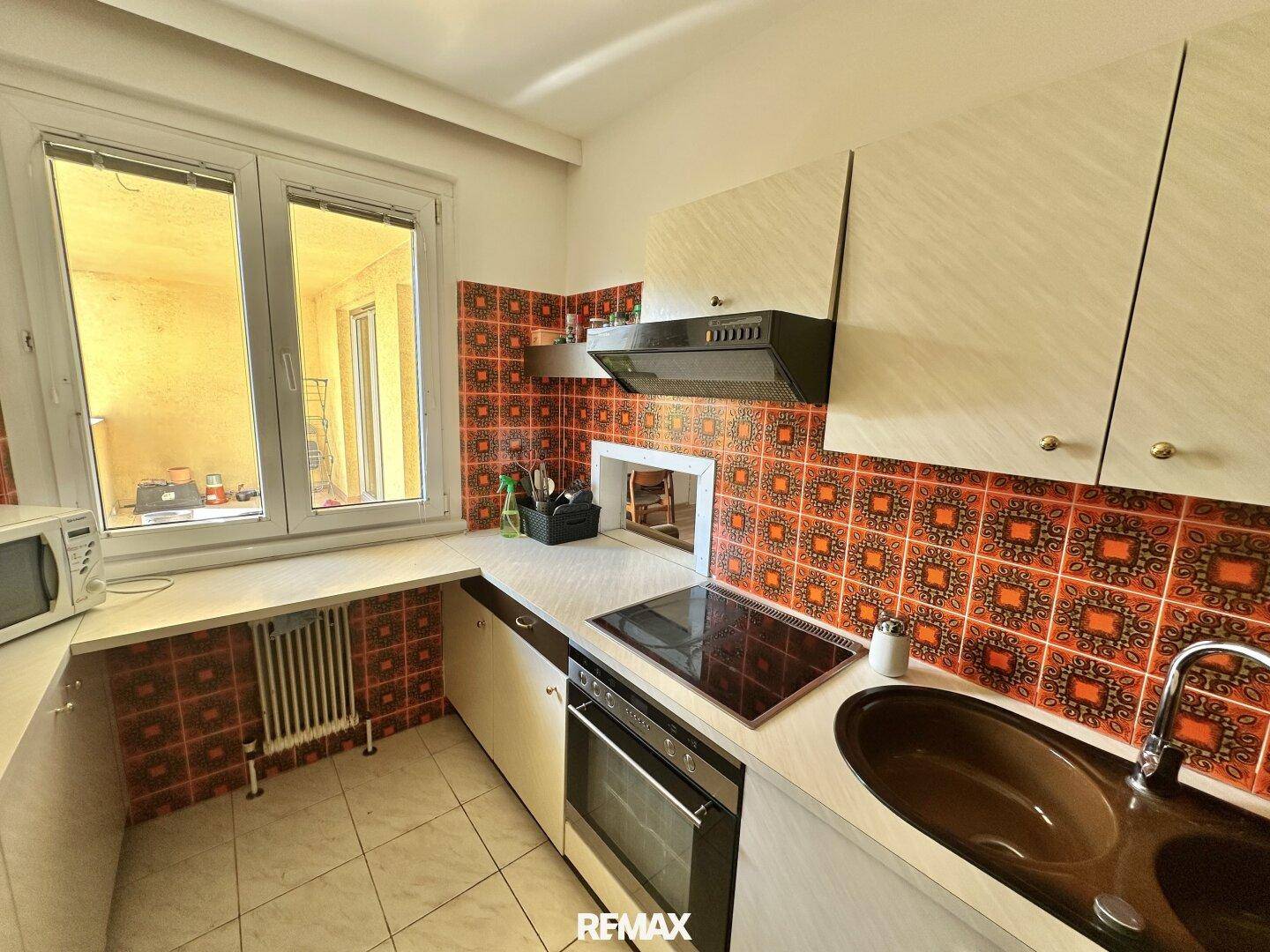 Küche mit Fliesenboden und roten Wandfliesen, ausgestattet mit Kochfeld, Backofen und Spüle unter dem Fenster.