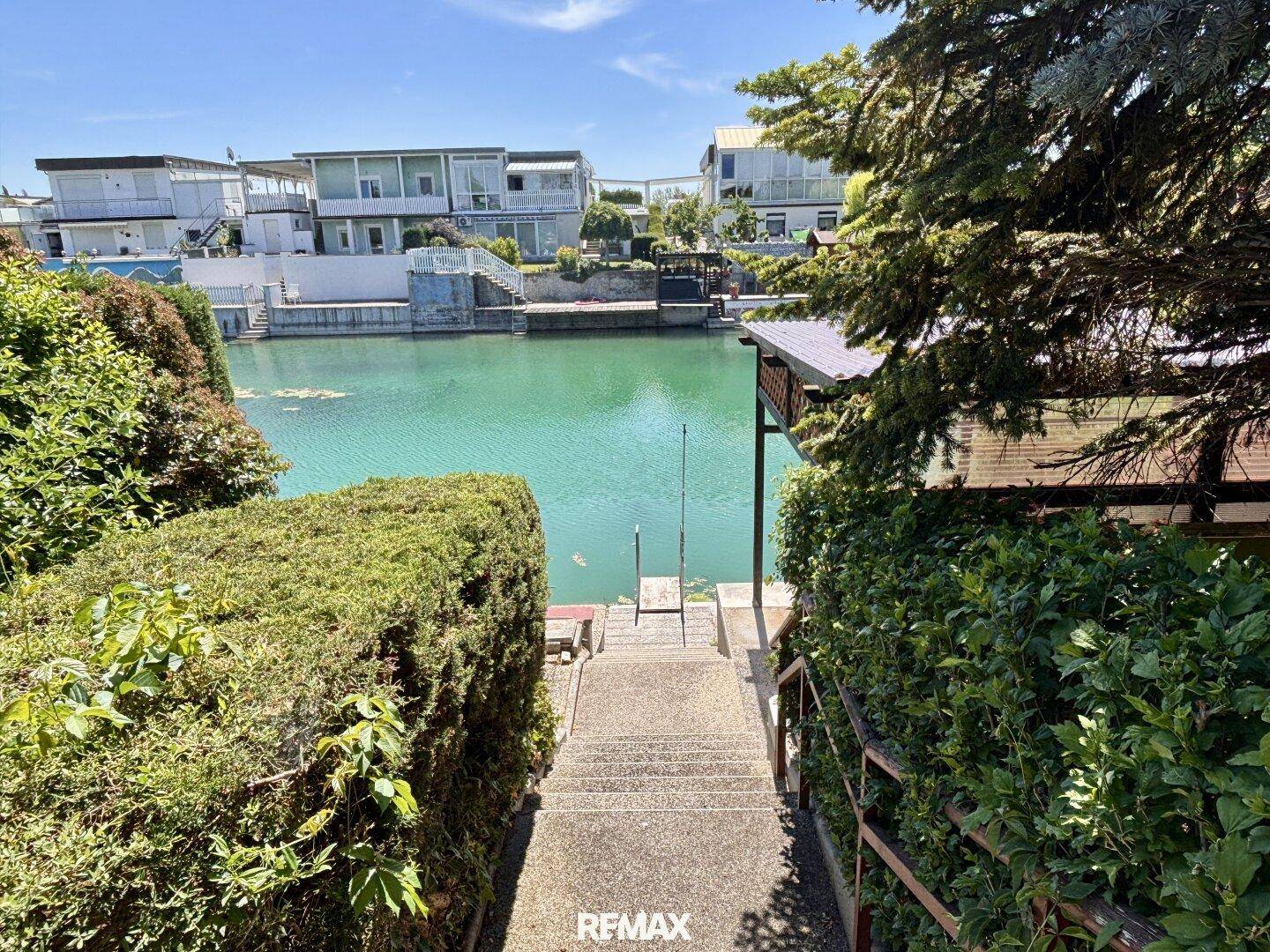 Blick von der Treppe durch dichte Hecken auf einen idyllischen See oder Kanal.