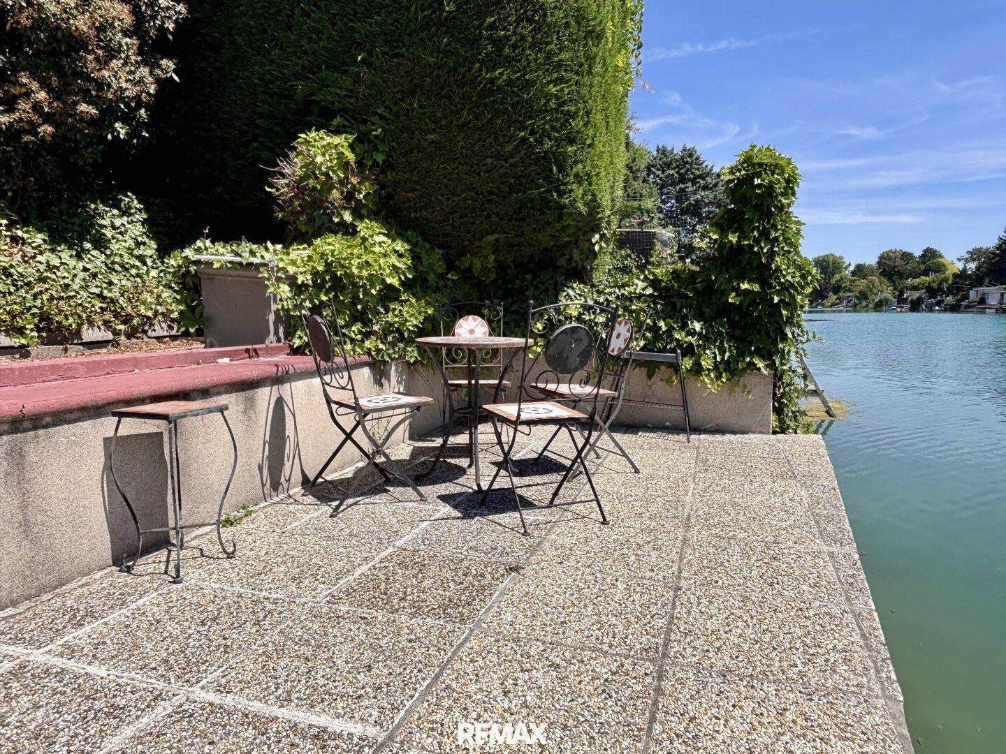 Gepflasterte Terrasse direkt am Wasser mit Gartenmöbeln und üppiger Vegetation.