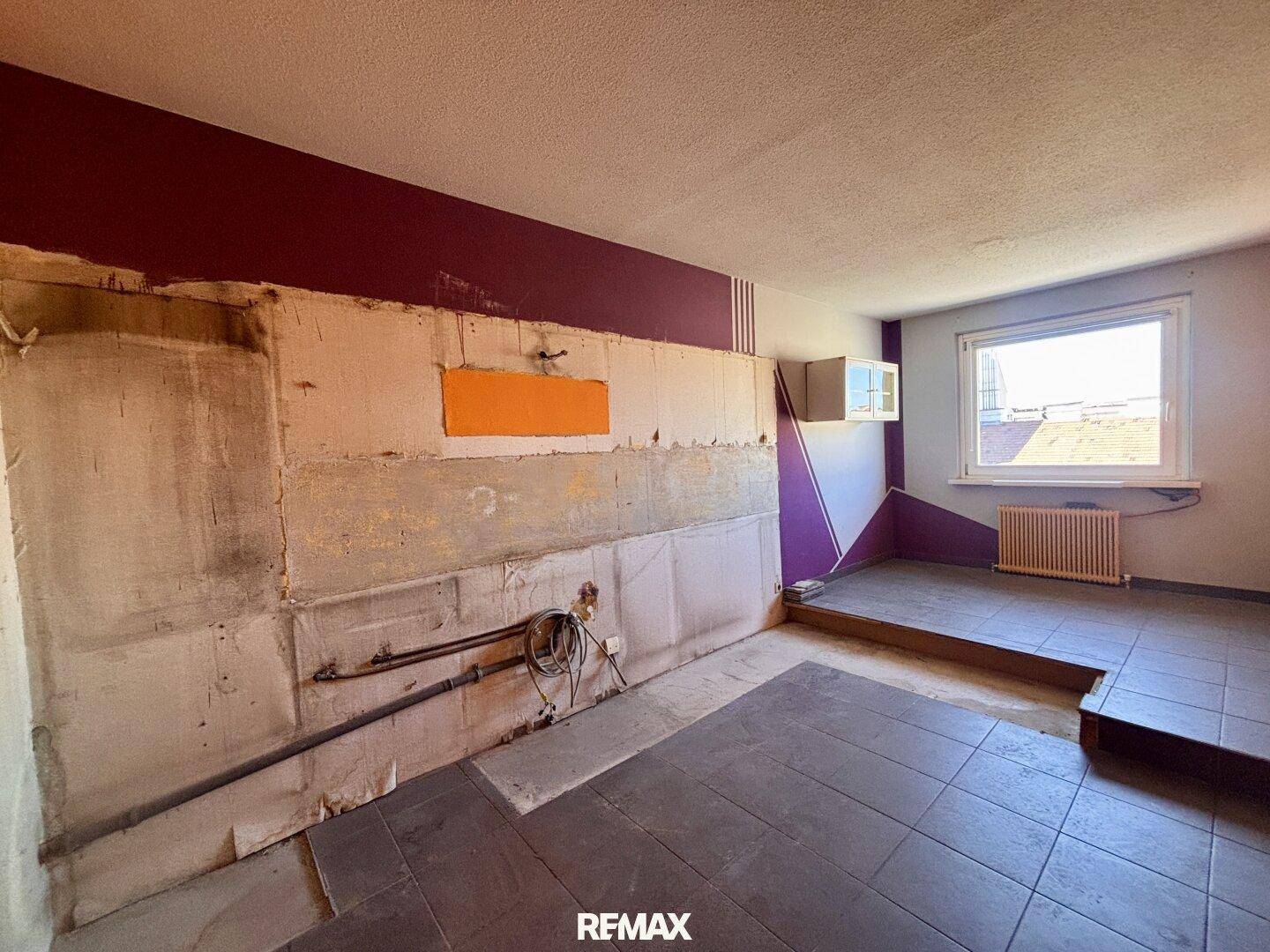 Renovierungsbedürftiger Raum mit freigelegter Wand, Fliesenboden und Spuren einer ehemaligen Küche.