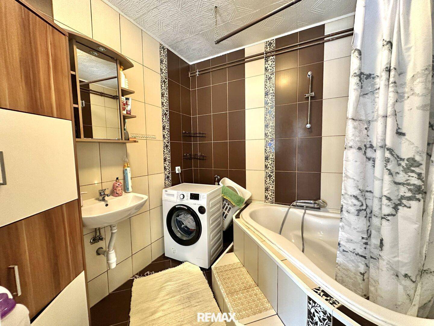Badezimmer mit Badewanne, Waschbecken und Waschmaschine, gefliest in Braun- und Beigetönen.