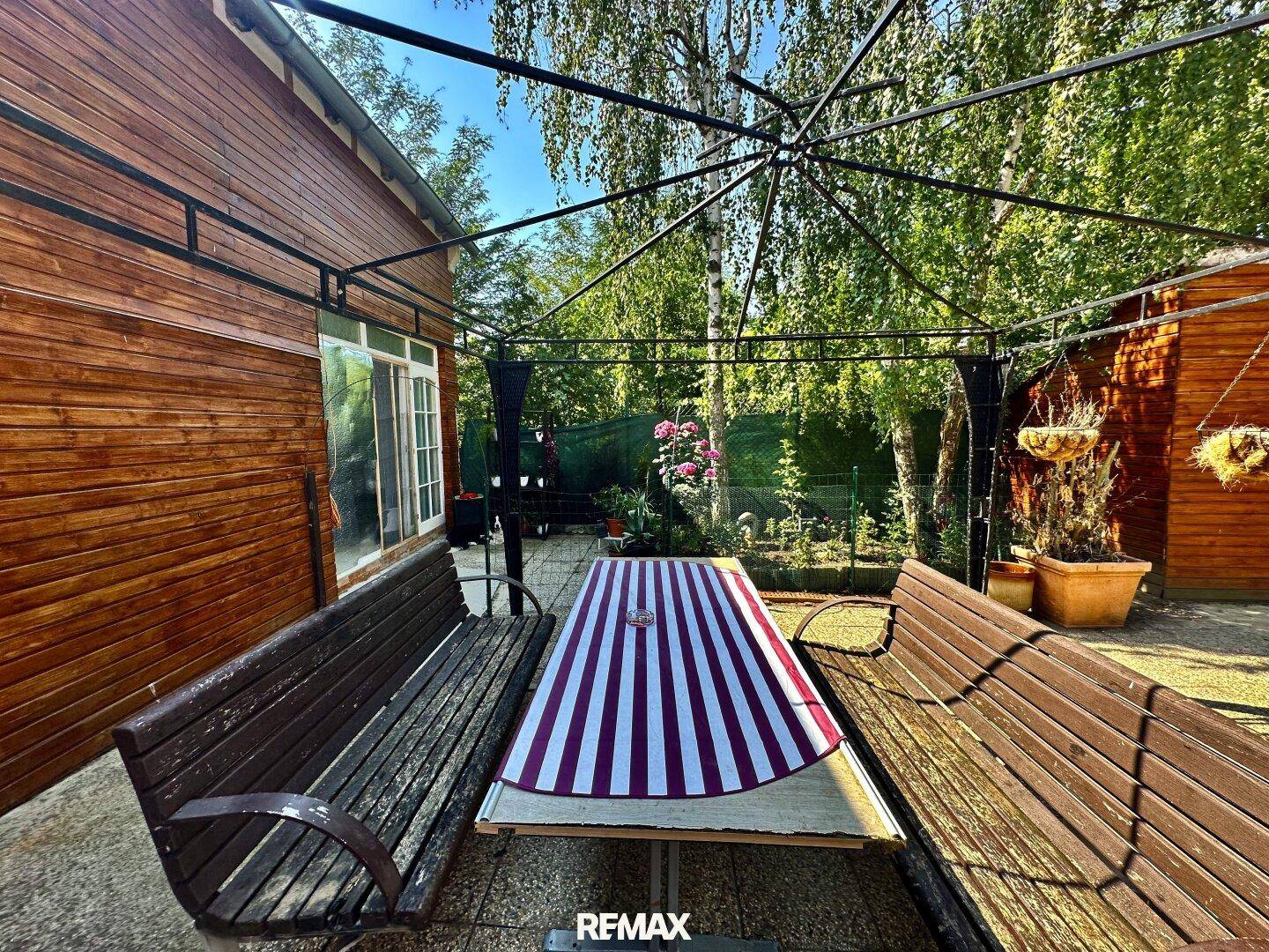 Gemütlicher Essbereich im Freien unter einer Pergola, umgeben von Bäumen und Pflanzen.