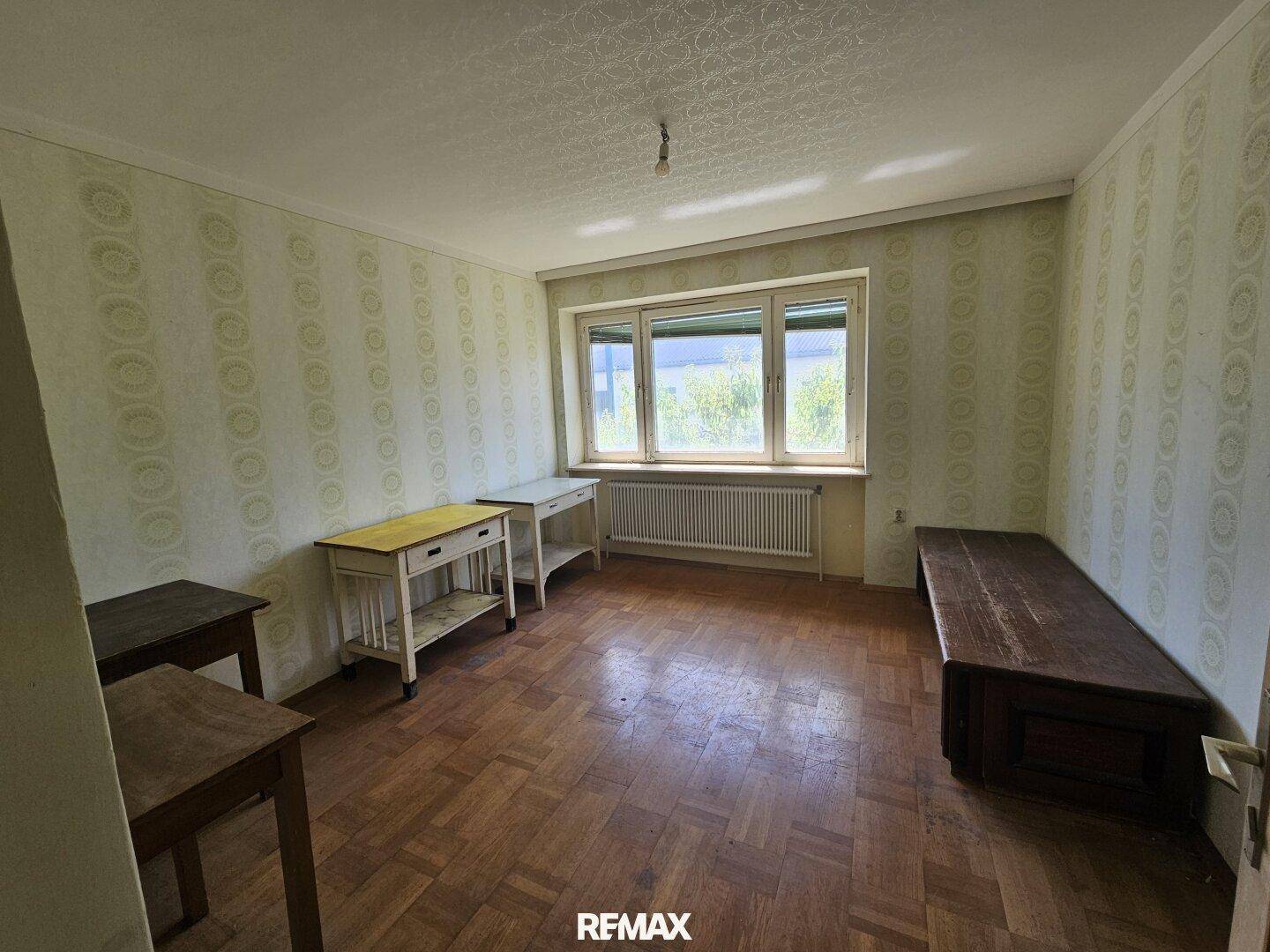 Geräumiges Zimmer mit Parkettboden, gemusterter Tapete und einfachen Holzmöbeln.