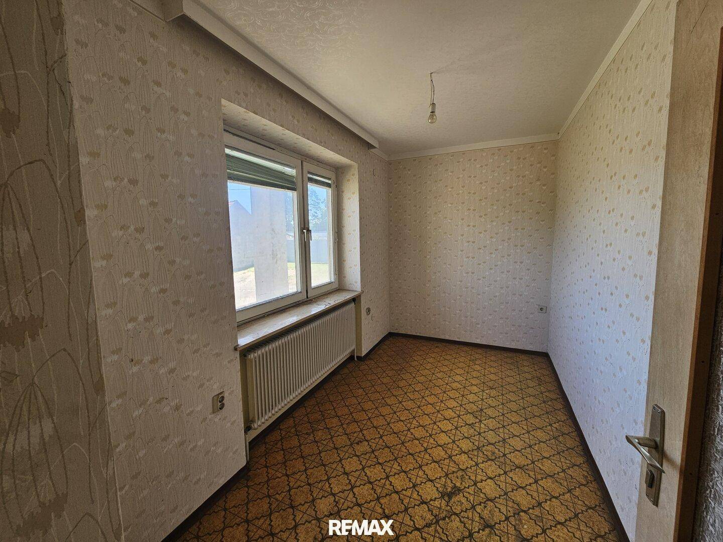 Zimmer mit gemusterter Tapete, Heizkörper und Fenster mit Blick nach draußen.