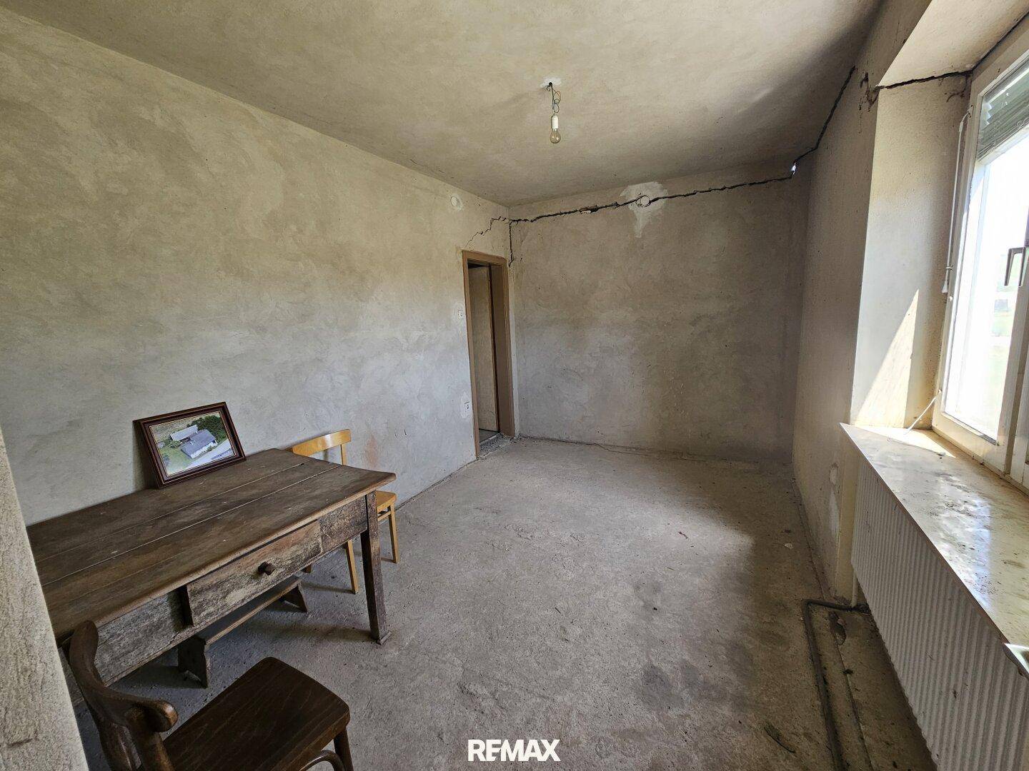 Unrenoviertes Zimmer mit Putzwänden, Holztisch und Fenster mit Blick ins Grüne.