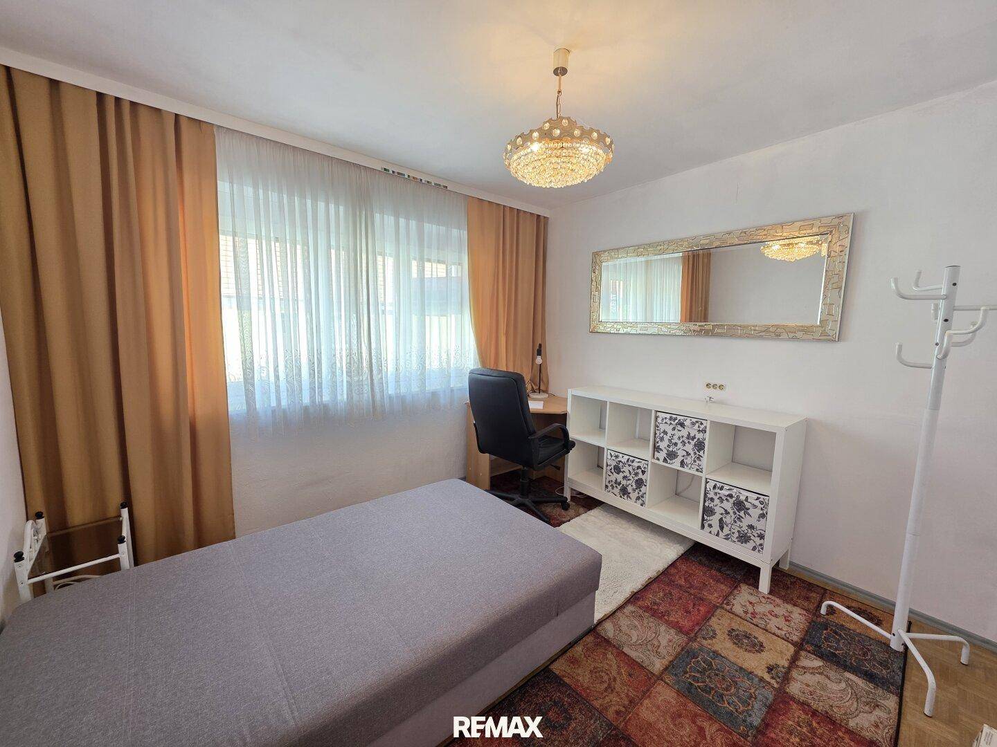 Helles Schlafzimmer mit großem Fenster, Teppichboden und einem kleinen Schreibtischbereich.