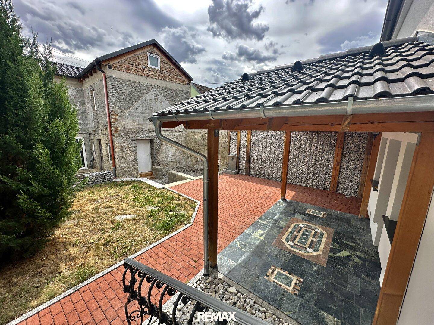 Überdachte Terrasse mit Ziegelpflaster und Blick auf ein älteres Nebengebäude.