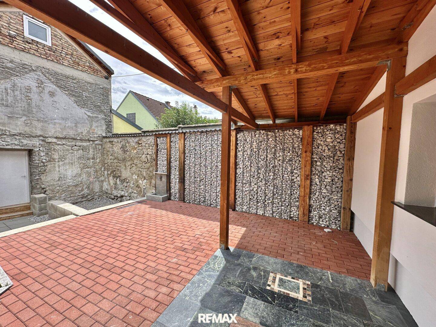 Überdachter Terrassenbereich mit Ziegelpflaster, Gabionenwänden und Blick auf ein altes Gebäude.
