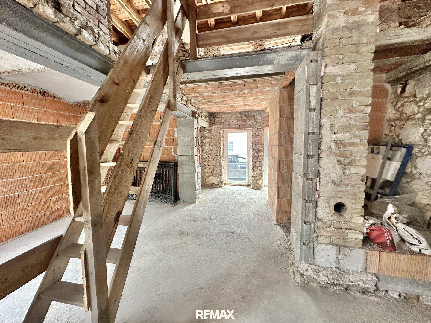 Innenraum im Rohbau mit freiliegenden Ziegelwänden, Holztreppe und sichtbaren Deckenbalken.