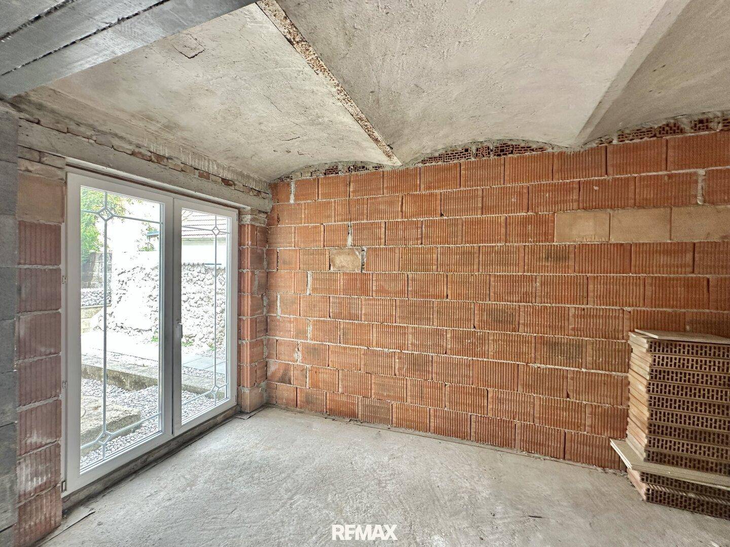 Rohbauzimmer mit unverputzten Ziegelwänden, Betonboden und großen Fenstern zum Garten.
